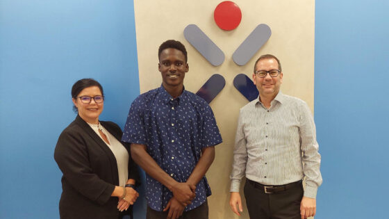 Drei Personen stehen vor einer blauen Wand mit einem Logo. Eine Frau in einem schwarzen Blazer und Brille lächelt. Ein Mann in einem blauen Hemd mit Mustern steht in der Mitte. Ein weiterer Mann in einem gestreiften Hemd steht rechts. Das Logo zeigt eine rote und blaue Grafik.