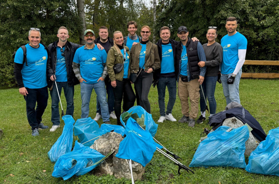 Eine Gruppe von zehn Personen steht auf einer Wiese, umgeben von Bäumen. Alle tragen blaue T-Shirts mit einem Logo. Einige halten Müllgreifer, während blaue Müllsäcke im Vordergrund liegen. Die Personen lächeln und posieren für das Foto, was auf eine umweltfreundliche Aktivität hindeutet.