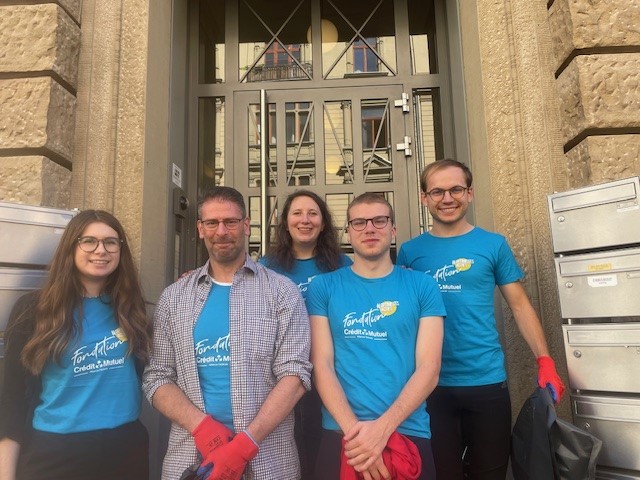 Fünf Personen stehen vor einem Gebäude, das einen Eingang mit einem metallenen Gitter hat. Sie tragen blaue T-Shirts mit einem Logo. Die Gruppe besteht aus zwei Frauen und drei Männern, wobei einer von ihnen eine Brille trägt. Alle lächeln und scheinen bereit zu sein, während sie rote Handschuhe tragen. Im Hintergrund sind Briefkästen sichtbar.