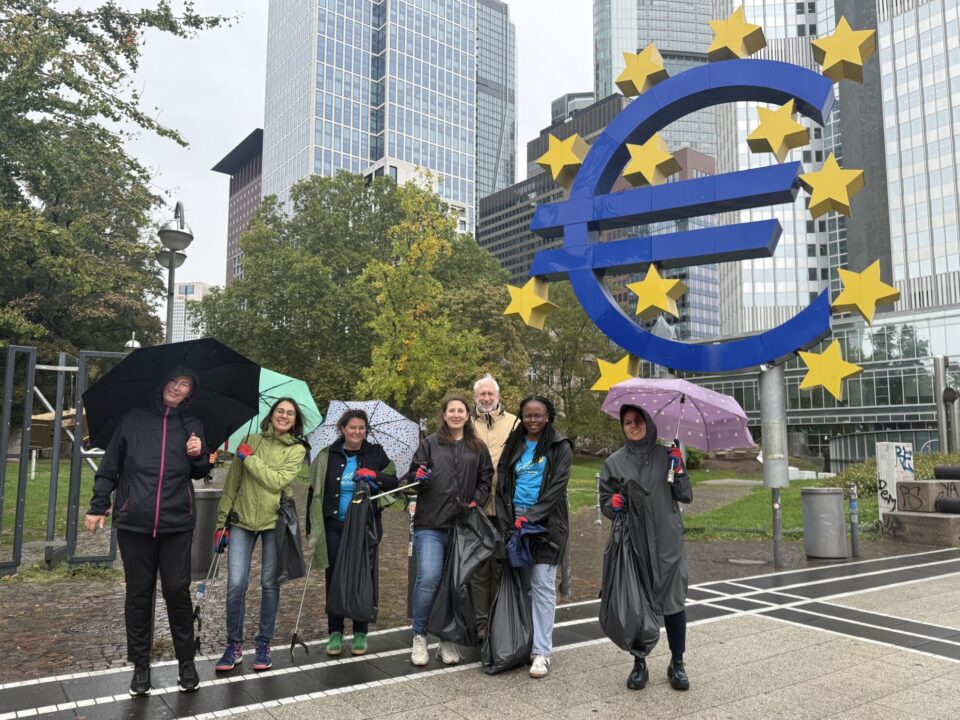 Eine Gruppe von sechs Personen steht unter Regenschirmen vor dem Euro-Symbol in Frankfurt. Es regnet leicht. Die Teilnehmer tragen wetterfeste Kleidung und halten Müllsäcke. Hinter ihnen sind moderne Gebäude sichtbar.
