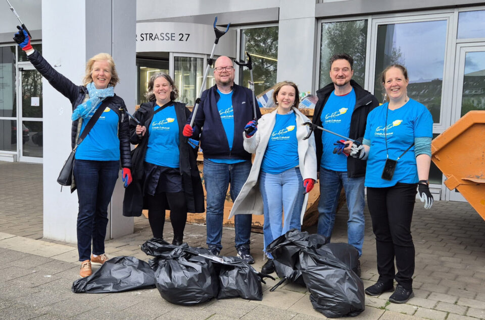 Eine Gruppe von sechs Personen steht vor einem Gebäude und lächelt. Sie tragen blaue T-Shirts mit einem Logo und halten Müllgreifer. Am Boden liegen mehrere schwarze Müllsäcke. Im Hintergrund sind Fenster und eine orangefarbene Abfallkippe zu sehen.