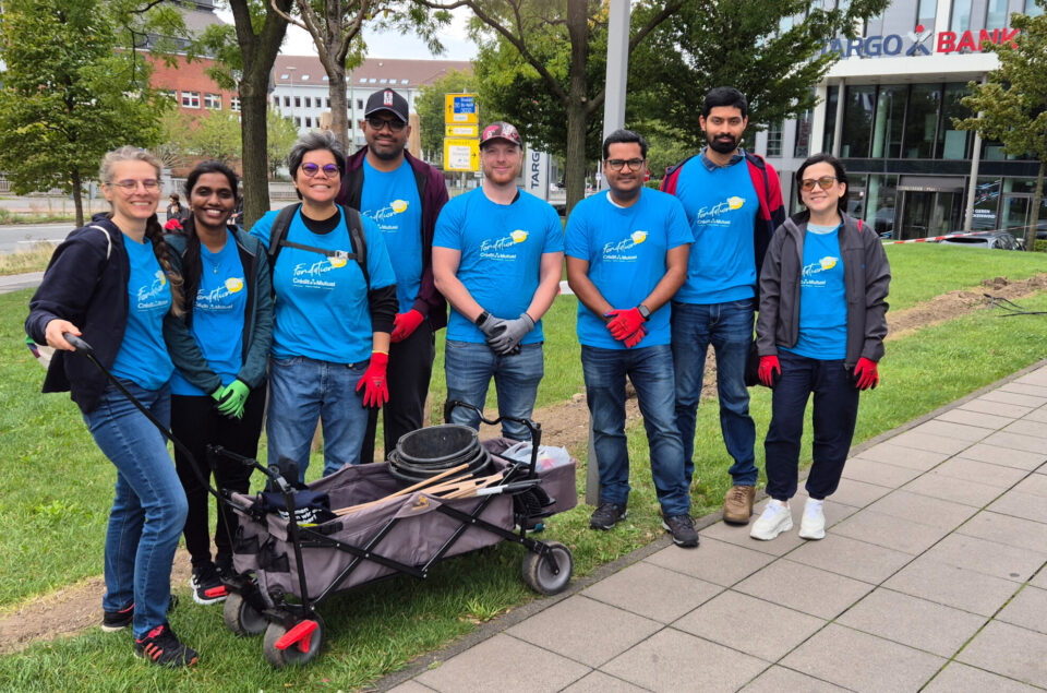 Eine Gruppe von acht Personen steht auf einer Rasenfläche vor einem Gebäude mit einem Logo der Bank. Sie tragen blaue T-Shirts mit einem gelben Logo und Handschuhe in verschiedenen Farben. Im Vordergrund befindet sich ein Kinderwagen mit Werkzeugen. Es ist ein bewölkter Tag.