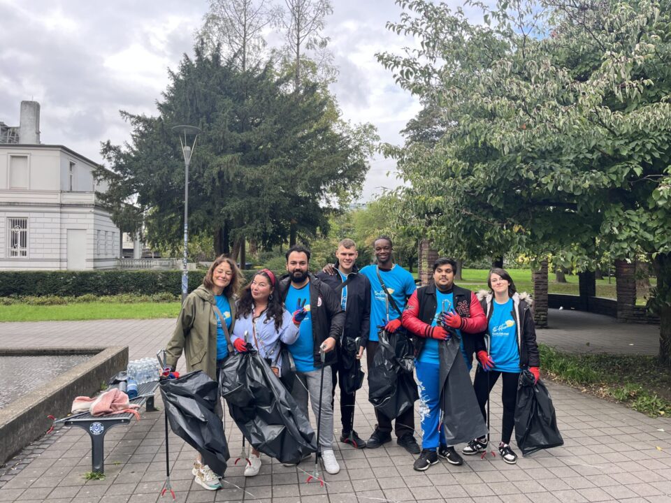 Eine Gruppe von sieben Menschen steht auf einem Gehweg in einem Park, umgeben von Bäumen und einem Gebäude im Hintergrund. Sie tragen blaue T-Shirts und haben Müllsäcke in der Hand. Die Stimmung ist freundlich, und einige Personen machen Peace-Zeichen. Im Hintergrund sind Bänke und Wasserflaschen sichtbar.