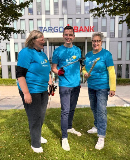 Drei Personen stehen auf einer grünen Wiese vor einem modernen Gebäude mit der Aufschrift 'TARGOBANK'. Alle tragen blaue T-Shirts mit einem Logo. Die Person in der Mitte hält eine Gartenschaufel, während die anderen zwei Werkzeuge in den Händen halten und lächeln.