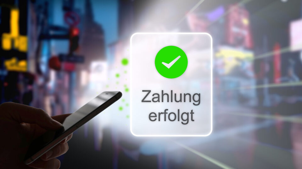 Eine Hand hält ein Smartphone, während auf dem Bildschirm ein grünes Häkchen und der Text "Zahlung erfolgt" angezeigt werden. Im Hintergrund sind unscharfe, beleuchtete Stadtlichter sichtbar.