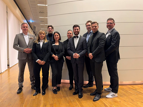 Eine Gruppe von neun Personen in formeller Kleidung steht in einem hellen Raum mit Holzfußboden. Sie posieren zusammen, einige mit Anzügen und andere in eleganter Kleidung.