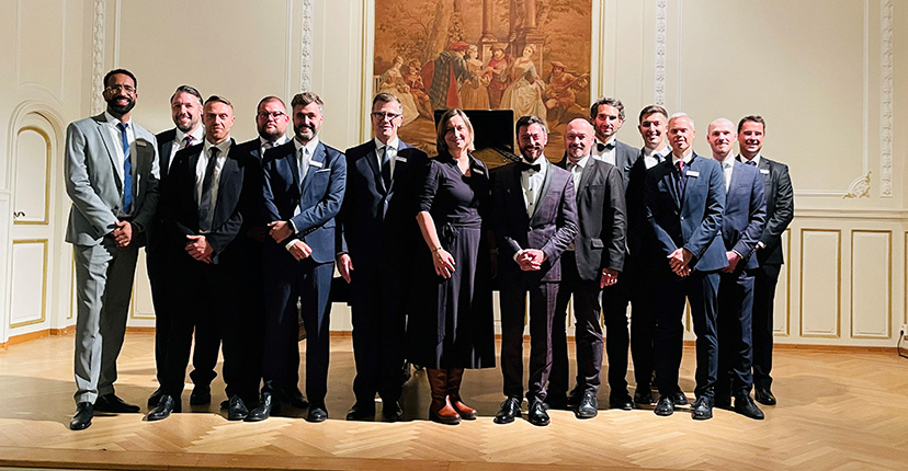 Eine Gruppe von zehn Männern und einer Frau steht auf einer Bühne, alle tragen formelle Kleidung. Im Hintergrund ist ein großes Wandgemälde zu sehen.