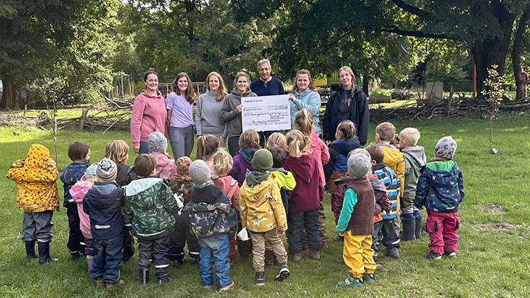 Eine Gruppe von Kindern in bunten Regenjacken steht im Freien und blickt auf Erwachsene, die ein großes Scheck halten. Die Szene spielt in einem grünen, baumreichen Bereich.