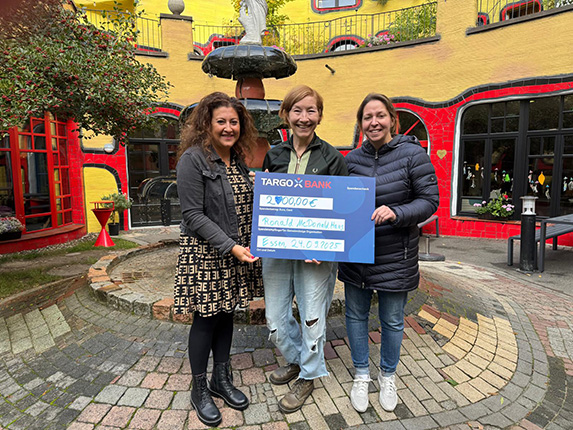 Drei Personen halten einen Scheck von TARGOBANK über 2.000,00 € in einer bunten Umgebung mit einem Brunnen im Hintergrund.