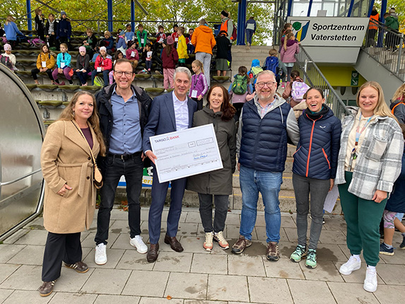 Eine Gruppe von Personen steht im Vordergrund und hält einen großen Scheck. Im Hintergrund sind Zuschauer auf Stufen sitzen, darunter Kinder in bunten Kleidern. Ein Schild mit der Aufschrift 'Sportzentrum Vaterstetten' ist sichtbar.