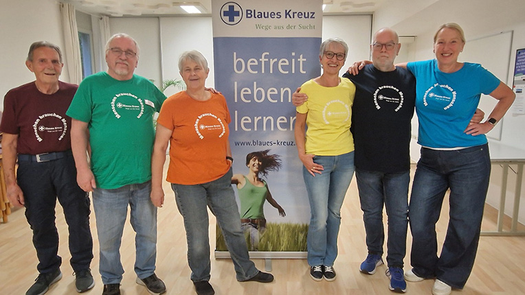 Sechs Personen in bunten T-Shirts stehen vor einem Banner des Blauen Kreuzes mit dem Text 'befreit leben lernen'.