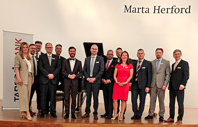 Eine Gruppe von zwölf Personen steht in formeller Kleidung vor einem Klavier in einem Raum mit dem Schriftzug 'Marta Herford'.