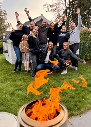 Eine Gruppe von zehn Menschen posiert fröhlich im Freien, während sie die Hände in die Luft heben. Im Vordergrund brennt ein Feuer in einer Metalltonne.