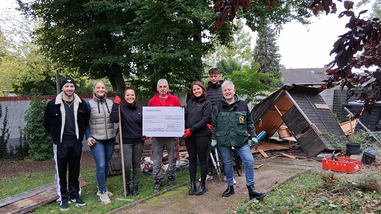 Sechs Personen stehen auf einem Weg in einem Garten. Sie halten ein großes Scheck-Schild mit dem Text "Spende für...". Im Hintergrund ist eine beschädigte Gartenhütte zu sehen, umgeben von Bäumen und Sträuchern.