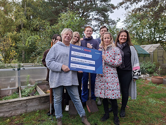 Eine Gruppe von Personen steht im Freien in einem Garten. Sie halten ein großes Schecksymbol mit dem Text: '40.000,00 €' und 'Schulförderung für die Stiftung'. Im Hintergrund sind Pflanzenbeete und ein Gewächshaus sichtbar.