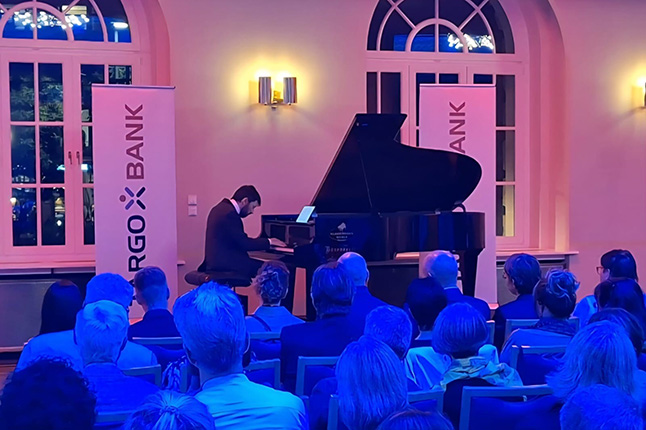 Ein Pianist spielt an einem Flügel während einer Veranstaltung, umgeben von einem Publikum in einem beleuchteten Raum mit Banner von der Cargo Bank.