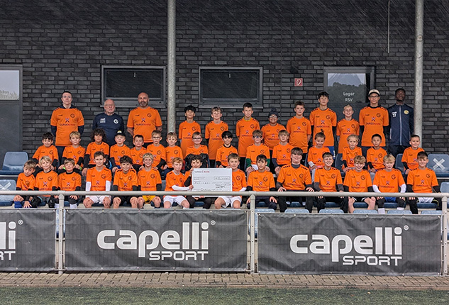 Eine Gruppe von 50 Kindern in orangefarbenen Trikots steht auf einer Sportanlage. Im Hintergrund sind Trainer und Betreuer sichtbar. Ein Banner mit dem Logo 'capelli SPORT' ist zu sehen.