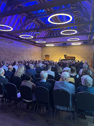 Eine große Menschenmenge sitzt in einem Raum mit Holzbalkendecke und blauen Lichtringen. Auf der Bühne steht ein Klavier.