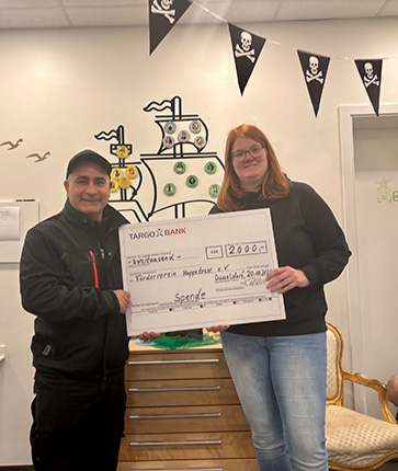 Zwei Personen halten einen großen Scheck mit einem Betrag von 2000 Euro. Im Hintergrund sind piratenartige Dekorationen zu sehen.