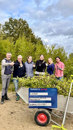 Sechs Personen stehen vor einem Schubkarren, der mit Pflanzen gefüllt ist. Sie zeigen Daumen hoch. Im Hintergrund sind Bäume und ein bewölkter Himmel zu sehen. Auf dem Schubkarren ist ein Schild mit der Aufschrift 'TARGOBANK' und einem Datum.