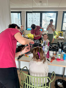 Eine Person mit langen Haaren sitzt auf einem Stuhl, während eine zweite Person mit rotem Shirt ihre Haare frisiert. Im Hintergrund sind weitere Personen und eine Vielzahl von Schönheitsprodukten auf dem Tisch sichtbar.