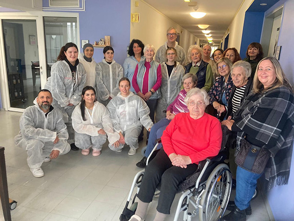 Eine Gruppe von Personen, die in einem Flur stehen und sitzen, trägt Schutzanzüge. Eine Person im Rollstuhl trägt ein rotes Oberteil. Die Umgebung ist hell mit weißen Wänden.
