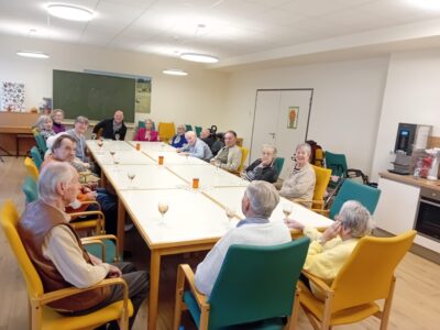 Eine Gruppe älterer Menschen sitzt an einem langen Tisch in einem hellen Raum. Auf dem Tisch stehen Gläser mit Getränken. Im Hintergrund sind Stühle in verschiedenen Farben und eine Kaffeemaschine sichtbar.