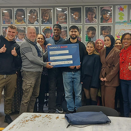 Eine Gruppe von Personen steht in einem Raum, in dem große Bilder an der Wand hängen. Sie halten einen großen Scheck von TARGOBANK über 2000 Euro. Einige Personen zeigen Daumen hoch.