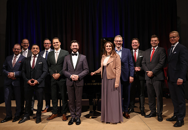 Eine Gruppe von Männern in Anzügen und einer Frau in einem eleganten Kleid stehen vor einem Klavier. Der Hintergrund ist dunkel.