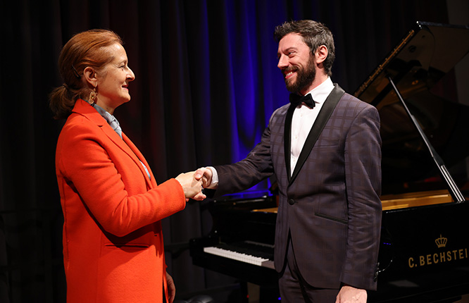 Eine Person in einem roten Mantel schüttelt einer anderen Person, die einen Smoking trägt, die Hand. Im Hintergrund steht ein Klavier.