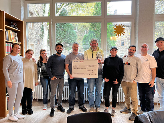 Eine Gruppe von Menschen steht zusammen in einem Raum mit großen Fenstern. In der Mitte hält jemand einen großen Scheck. Im Hintergrund sind Bücherregale und Fensterpflanzen zu sehen.