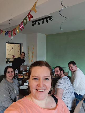 Eine Gruppe von fünf Personen sitzt an einem Tisch mit Tellern und Getränken. Im Hintergrund hängen bunte "HAPPY BIRTHDAY"-Bänder und Luftschlangen.