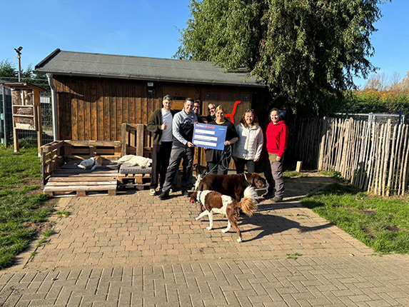 Eine Gruppe von fünf Personen steht vor einer Holzhütte im Freien, umgeben von einem eingezäunten Bereich. Sie halten ein großes Schild. Zwei Hunde sind sichtbar, einer ist braun und weiß, der andere ist schwarz und braun. Die Umgebung ist sonnig und grün.