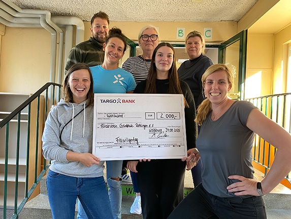 Eine Gruppe von Menschen steht zusammen und hält einen großen Scheck der TARGOBANK über 2.000 Euro. Der Scheck ist für die 'Förderverein Grüne Brücke e.V.' datiert auf den 29.12.2015. Die Szene ist im Innenbereich eines Gebäudes mit einer Treppe und einem Eingang.