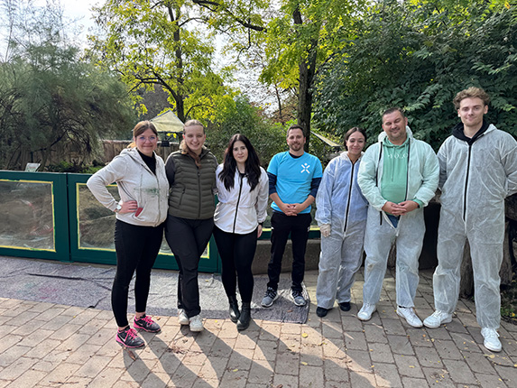 Eine Gruppe von sechs Personen steht im Freien, umgeben von Bäumen und einer grünen Wand. Einige tragen Schutzanzüge, andere lässige Kleidung.