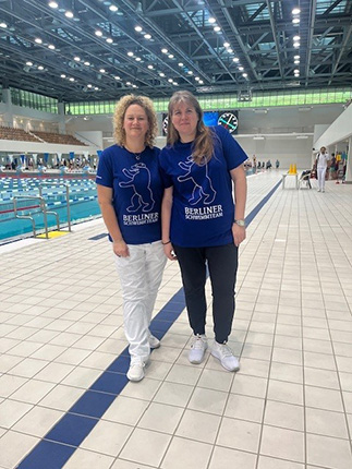 Zwei Personen stehen am Beckenrand eines Schwimmbads. Sie tragen blaue T-Shirts mit dem Aufdruck "BERLINER" und einer Grafik. Im Hintergrund sind Schwimmbahnen zu sehen.