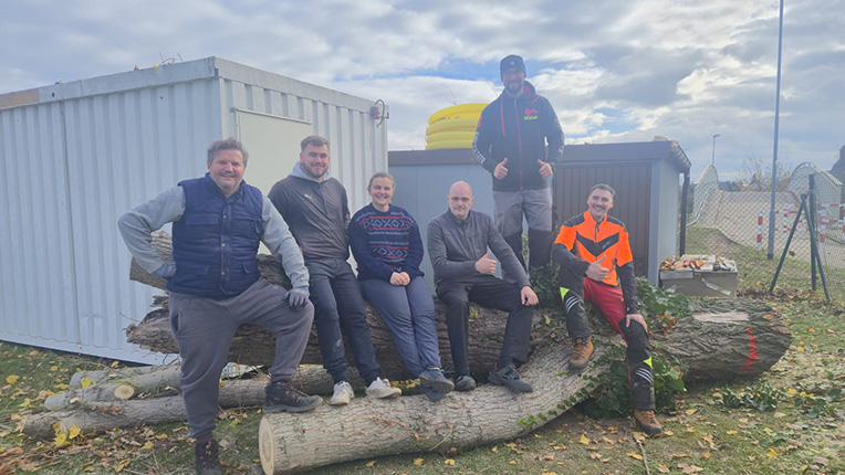 Sechs Personen sitzen auf einem Baumstamm, umgeben von einer grünen Wiese und einem Container im Hintergrund. Einige tragen Arbeitskleidung.