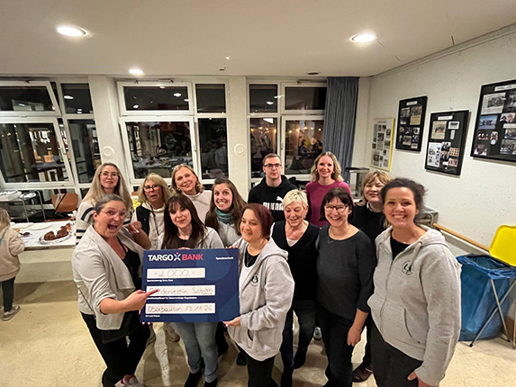Eine Gruppe von Personen hält ein großes Scheck über 2000 Euro, präsentiert von TARGOBANK. Im Hintergrund sind Fenster und Bilder an der Wand zu sehen.