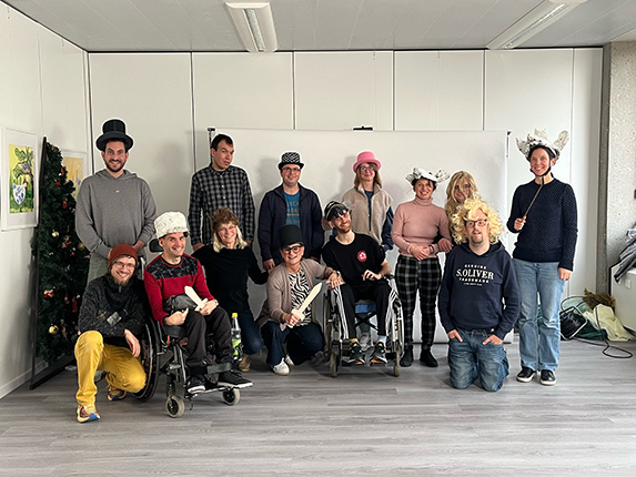 Eine Gruppe von zwölf Personen posiert in einem Raum mit einem weißen Hintergrund. Einige tragen verschiedene Hüte und Kostüme. Im Hintergrund steht ein Weihnachtsbaum.