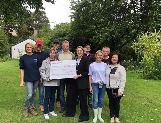 Eine Gruppe von Personen steht auf einer grünen Wiese, während eine Person einen großen Scheck hält. Im Hintergrund sind Bäume und ein Gebäude zu sehen.