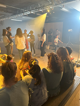 Eine Gruppe von Menschen steht in einem beleuchteten Raum, während andere Zuschauer auf einer Bank sitzen. Die Szene zeigt eine Mischung aus Erwachsenen und Kindern, die sich in einem kreativen Umfeld befinden, möglicherweise bei einer Aufführung oder Probe.