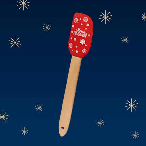 Ein roter Silikonspatel mit dem Aufdruck "Frohe Weihnachten" und Schneeflocken, auf einem blauen Hintergrund mit Sternen.