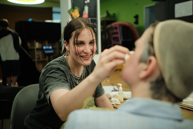 Eine Person, die eine andere Person mit Farben oder Make-up an einem Tisch bearbeitet, umgeben von verschiedenen Gegenständen.