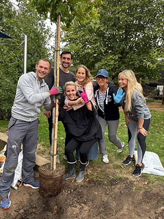 Eine Gruppe von Personen pflanzt einen Baum im Freien. Einige halten die Baumstamm, während andere helfen, ihn in die Erde zu setzen. Die Umgebung ist grün und es gibt eine Gartenlandschaft.