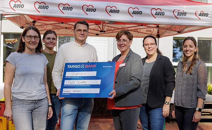 Eine Gruppe von Personen steht unter einem Zelt mit dem Logo der AWO. Eine Person hält einen großen Scheck der TARGOBANK über 2.000 €. Der Scheck ist beschriftet mit "AWO Diakonie" und dem Datum.