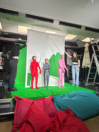 Vier Personen stehen auf einer grünen Bühne, zwei in roten Anzügen, einer in grau und einer in pink. Im Hintergrund ist ein grünes Tuch und eine Leiter sichtbar.