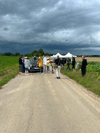 Eine Gruppe von Menschen versammelt sich auf einer Straße, umgeben von Feldern. Einige tragen helle Kleidung, während andere dunkle Kleidung tragen. Im Hintergrund ist ein Zelt sichtbar, und es gibt Kameras und Technik. Der Himmel ist bewölkt und droht mit Regen.