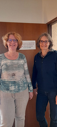 Zwei Frauen stehen nebeneinander vor einer Holztür mit einem Schild, auf dem "Alzheimer Gesellschaft" steht. Die linke Frau trägt einen blauen Pullover mit einem Muster und graue Hosen, während die rechte Frau einen dunklen Pullover anhat und Jeans trägt.