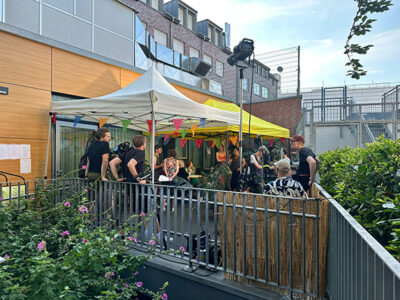 Eine Gruppe von Menschen steht unter zwei Zelten, umgeben von Pflanzen. Einige haben Kameras. Im Hintergrund sieht man ein Gebäude mit Fenstern.