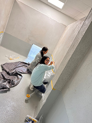 Zwei Frauen, eine in einem grauen Overall und die andere in einem blauen Sweatshirt, streichen eine Wand in einem Raum. Der Boden ist grau und es liegt abdeckendes Material auf dem Boden. Eine der Frauen lächelt in die Kamera.